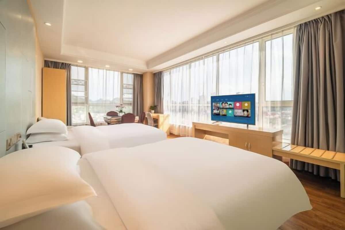 A hotel room with two beds, a TV, a desk, and large windows.