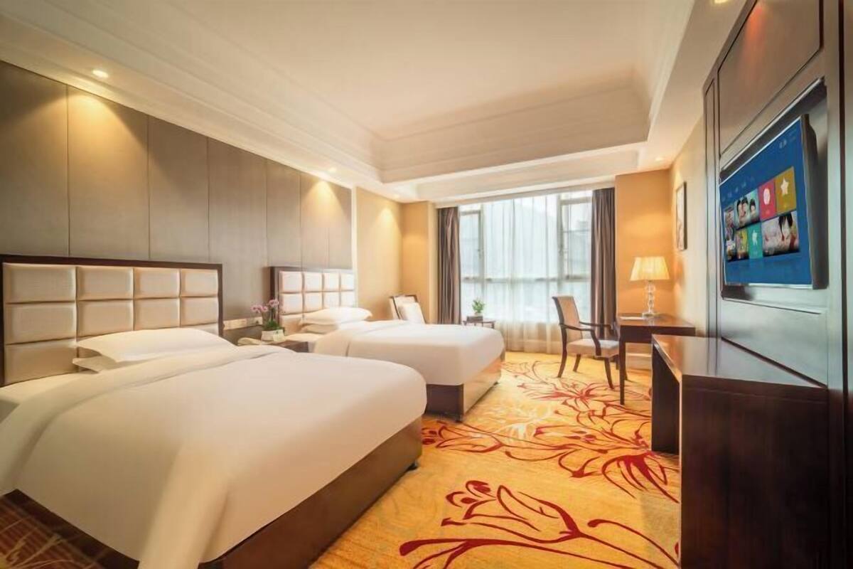 A hotel room with two beds, a desk, a chair, a TV, and a large window.