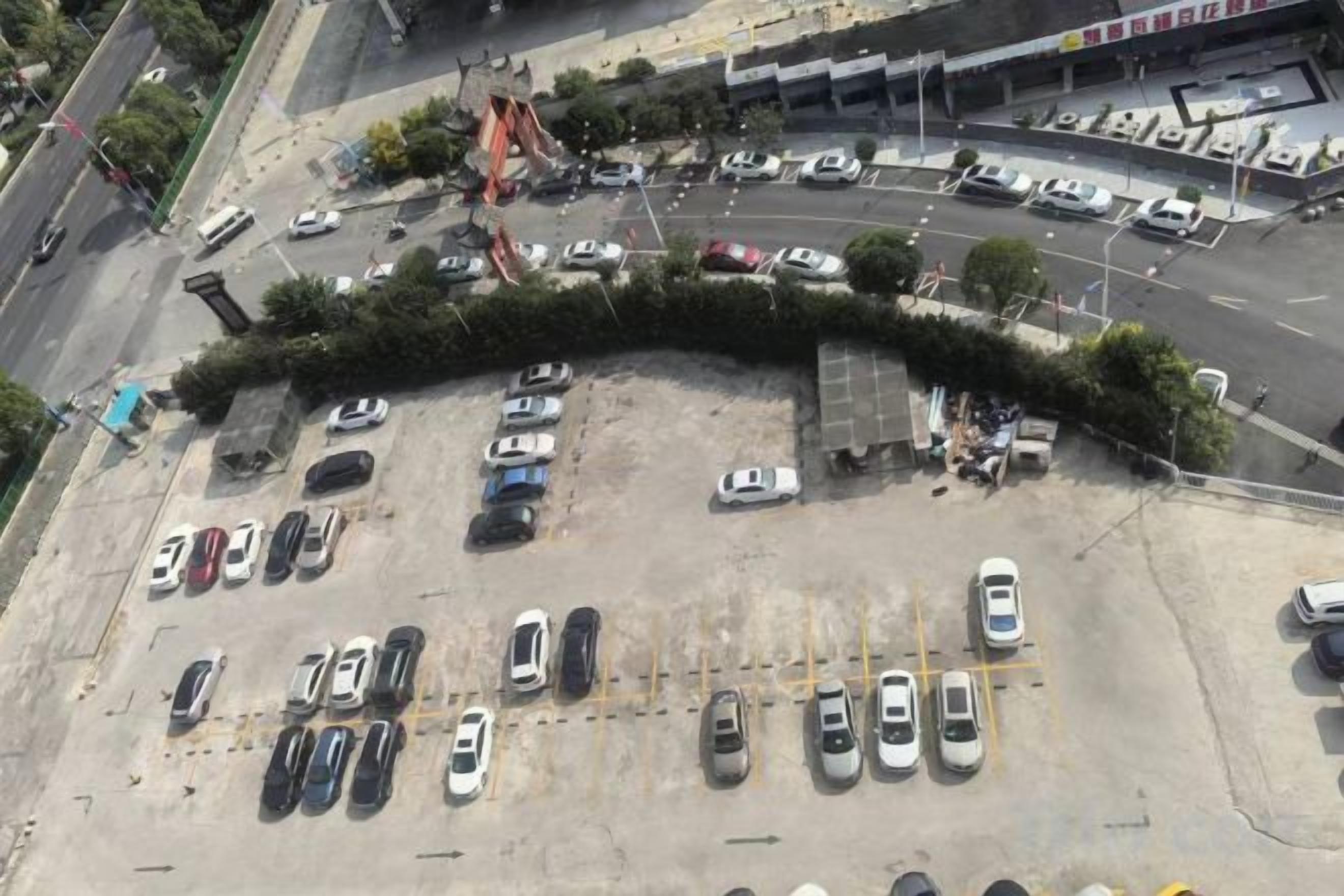 Eine Vogelperspektive auf einen Parkplatz mit mehreren Reihen geparkter Autos, einer gewundenen Grünhecke und einem Gebäude im Hintergrund.