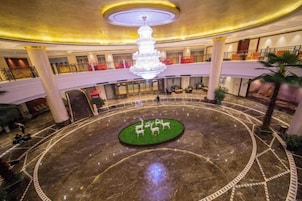 Um saguão de hotel com área circular central e uma fonte, cercado por um chão de mármore com padrões geométricos.