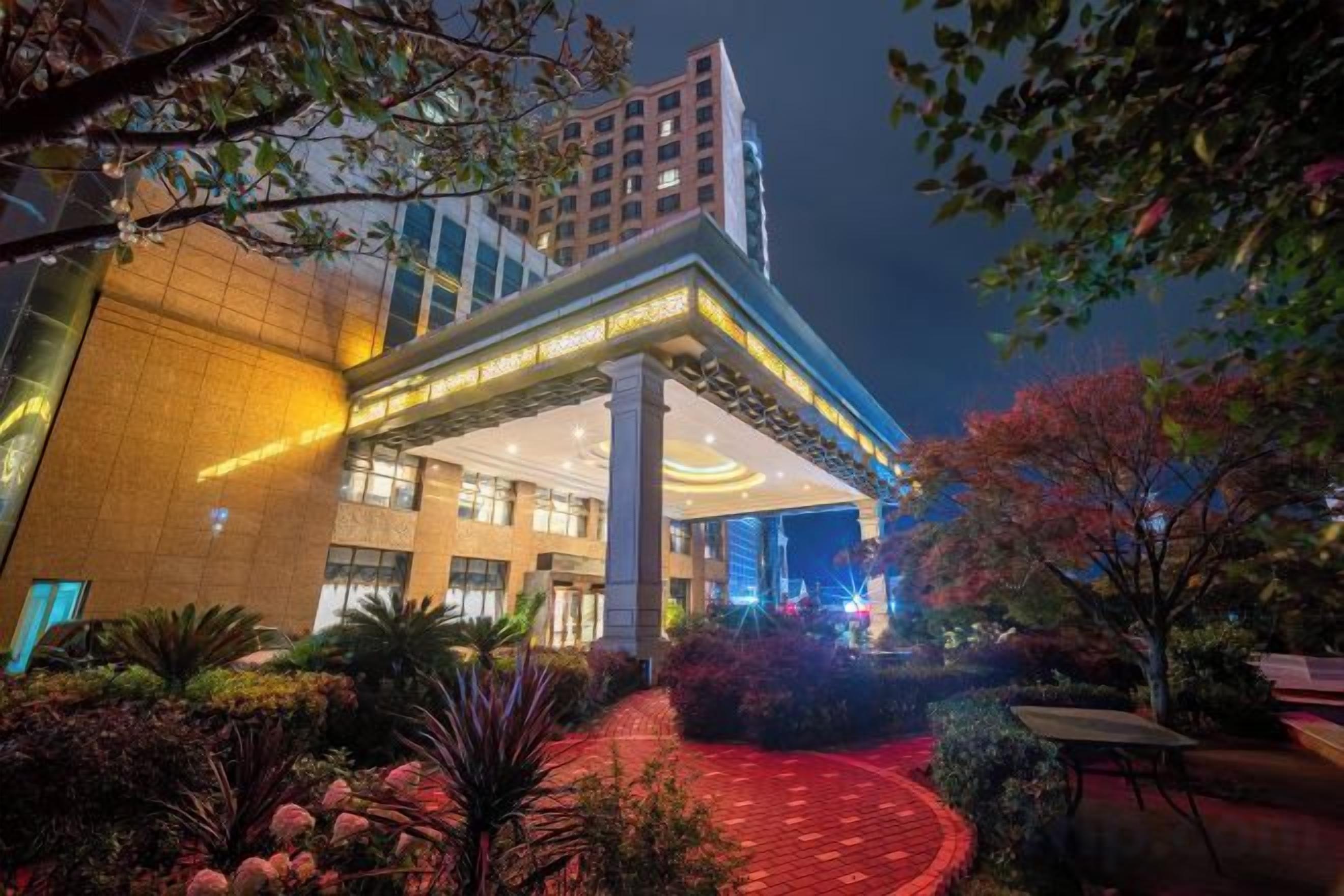 Un hotel con diseño moderno, que cuenta con una entrada destacada con galería techada, fachada iluminada y un jardín ajardinado.