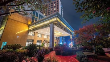 Un hotel con diseño moderno, que cuenta con una entrada destacada con galería techada, fachada iluminada y un jardín ajardinado.