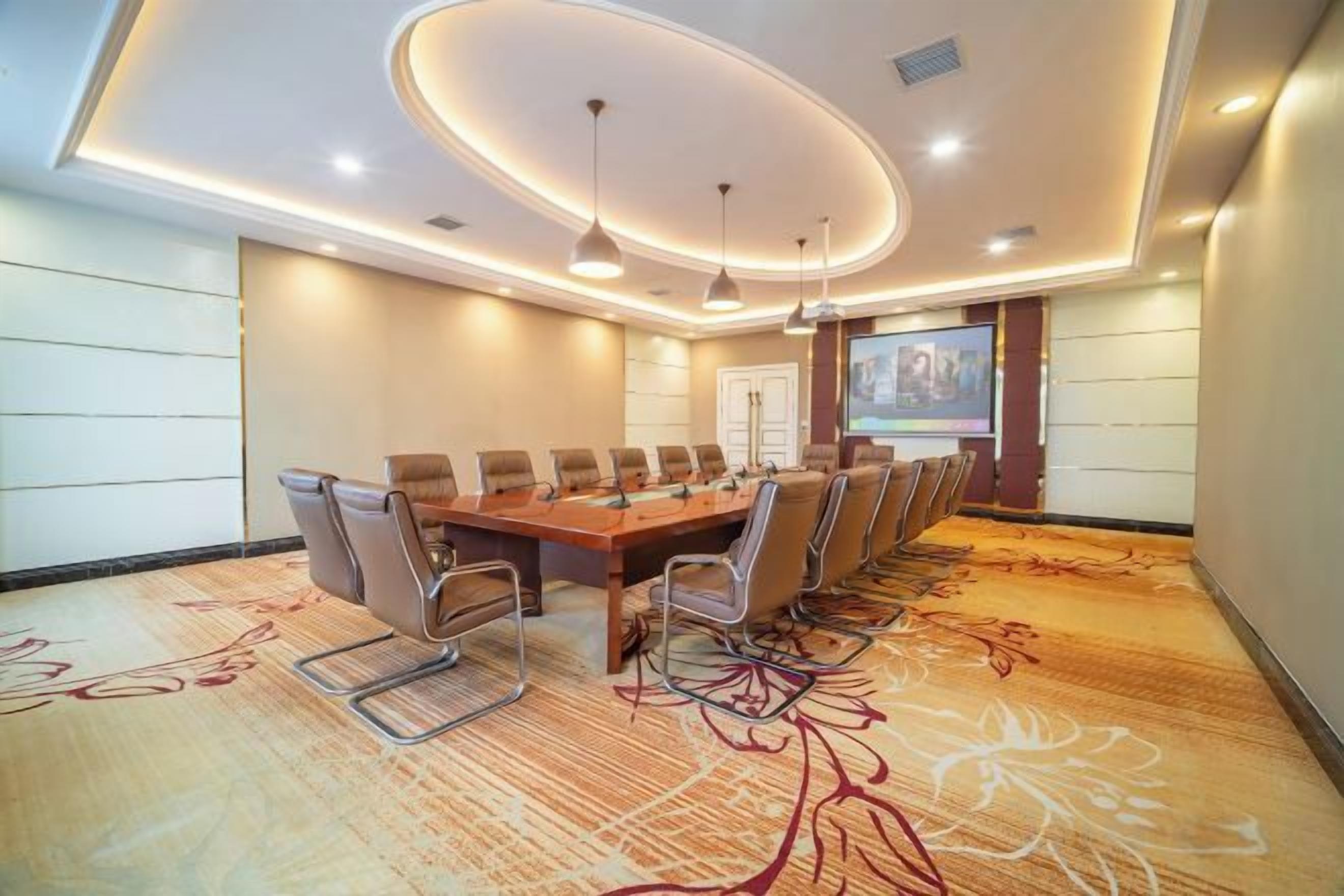 Een vergaderzaal met een lange tafel, stoelen, een projectiescherm en een vloerkleed met een bloemendesign.