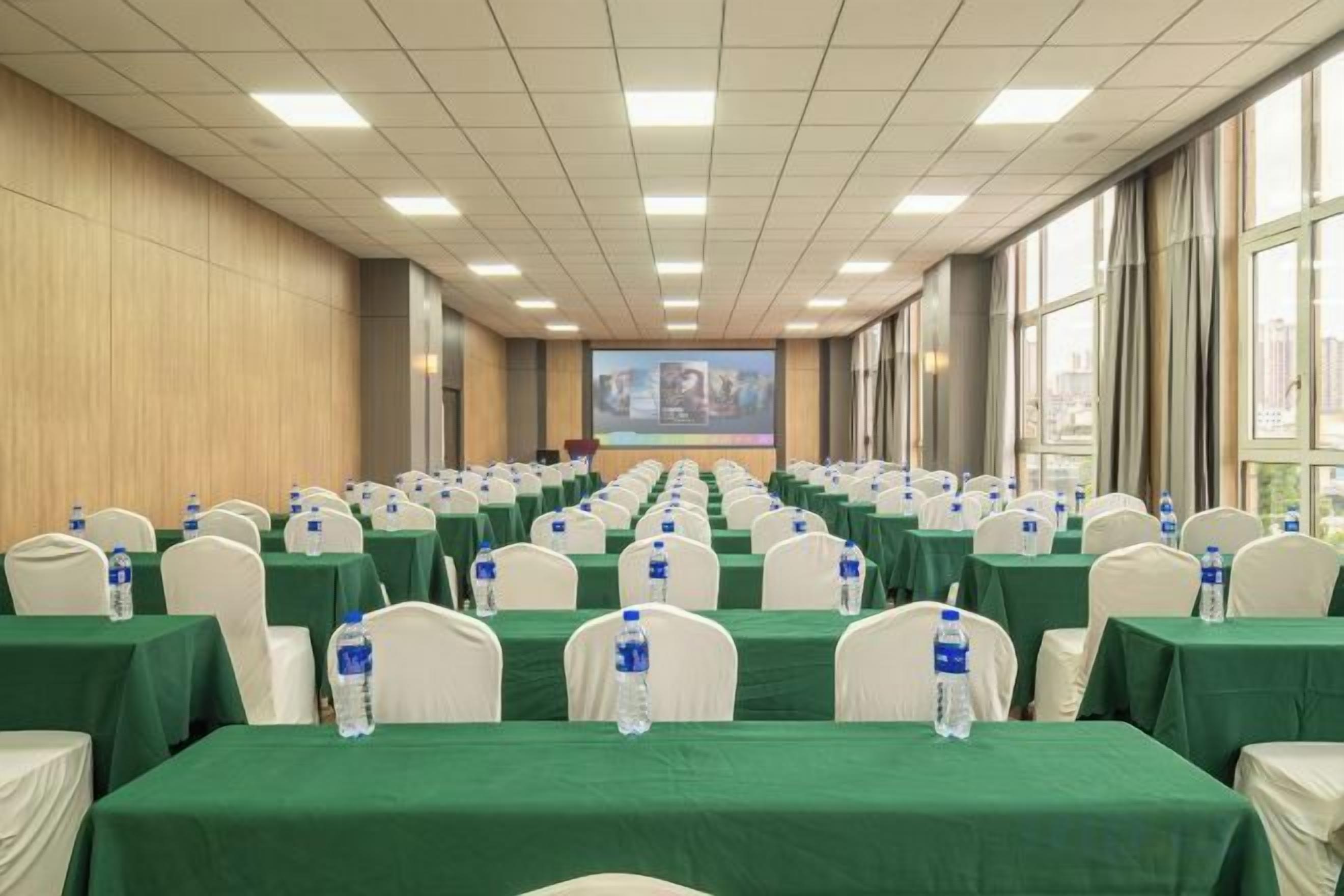 A conference room with rows of tables, chairs, and water bottles, equipped with a large screen at the front.