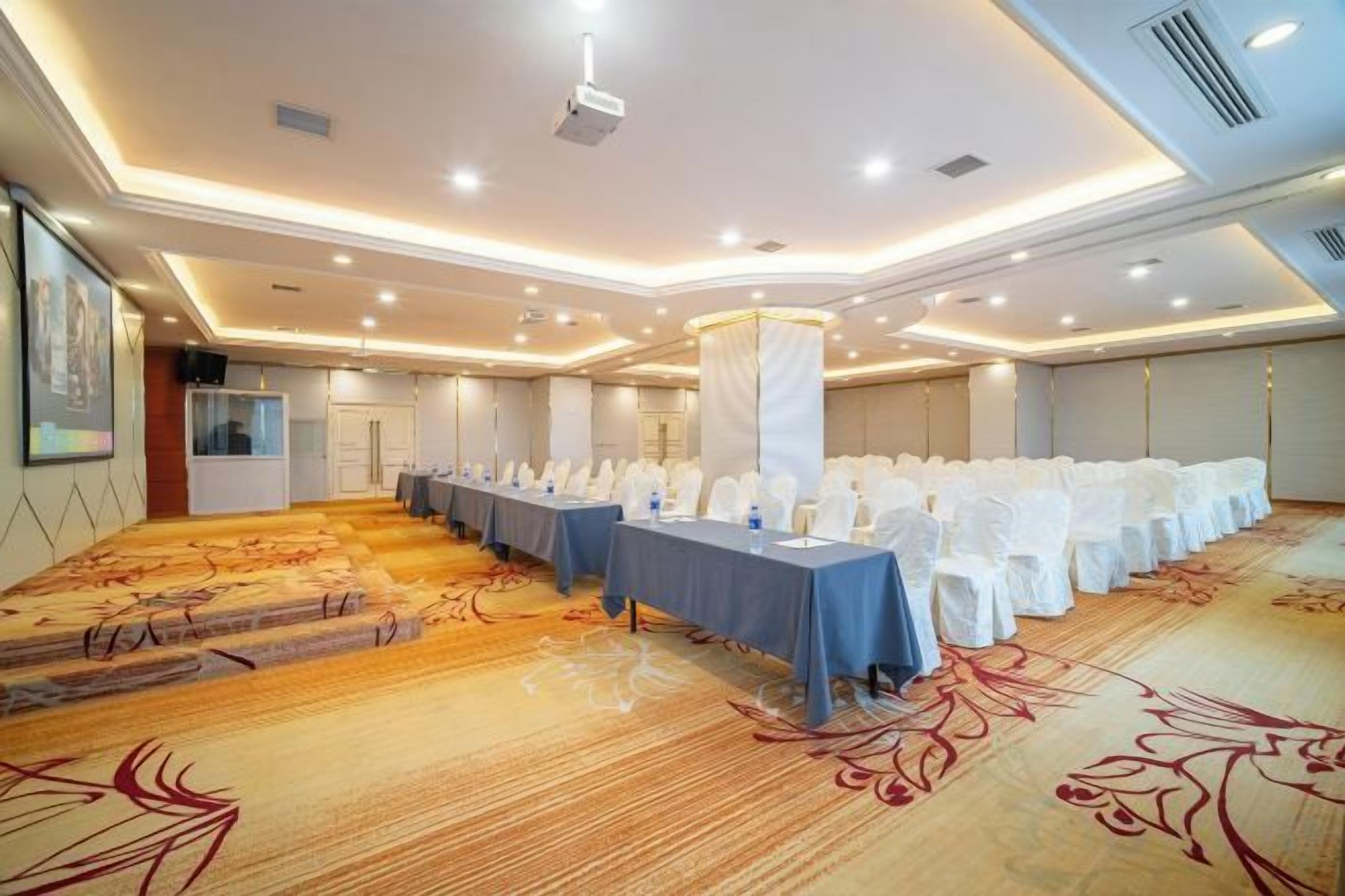 Un salón de conferencias con filas de mesas dispuestas para una reunión, suelo alfombrado e iluminación empotrada en el techo.