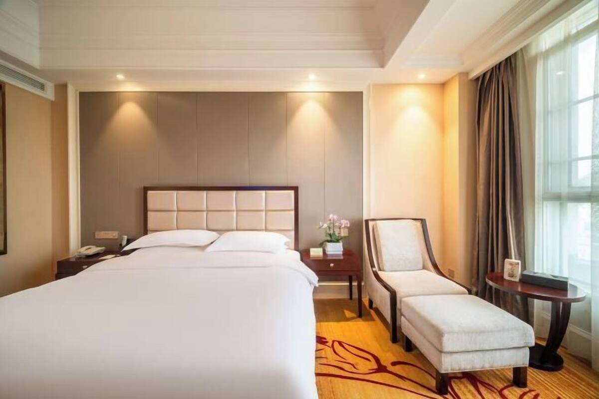 A hotel room with a large bed, a chair, a small table, and a window with curtains.