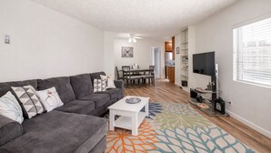A modern living room with a grey sofa, a white coffee table, and a colorful rug. There is a dining area with a table and chairs, and a kitchen in the background. - Stay in the Heart of Hillcrest! San Diego (San Diego)