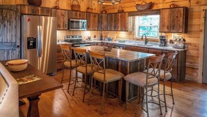 A rustic cabin-style kitchen with wooden walls, a central island with bar stools, a refrigerator, stove, and a window overlooking greenery. - Pinhoti Ridge Retreat Cabin 2, Skyline (Talladega)