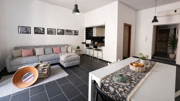 Un salon moderne avec un canapé gris, une table à manger agrémentée d’un chemin de table à motifs, et un coin cuisine doté de meubles blancs et d’un plan de travail noir.
