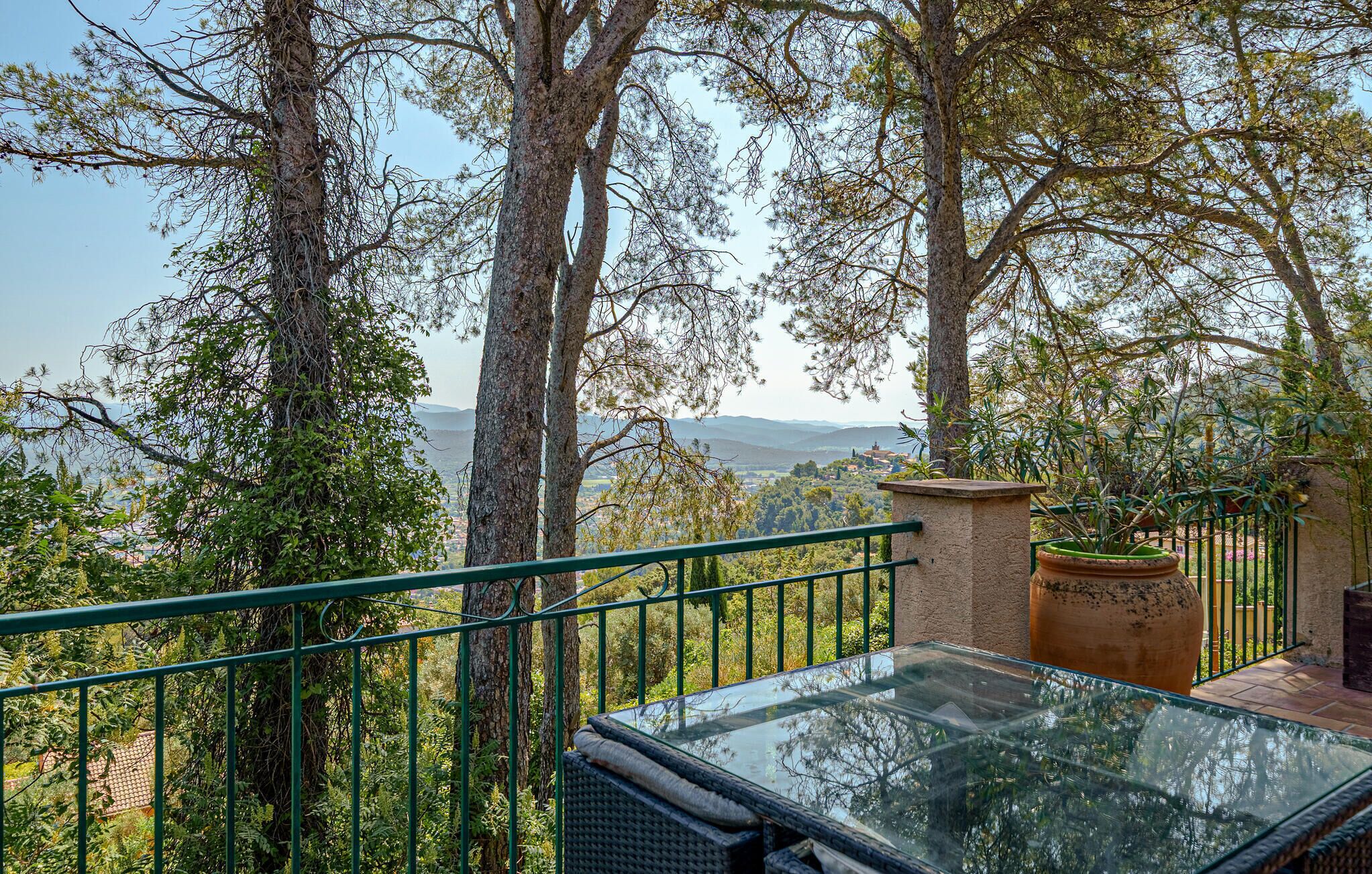 A balcony with a glass table, surrounded by trees and a view of a distant landscape.