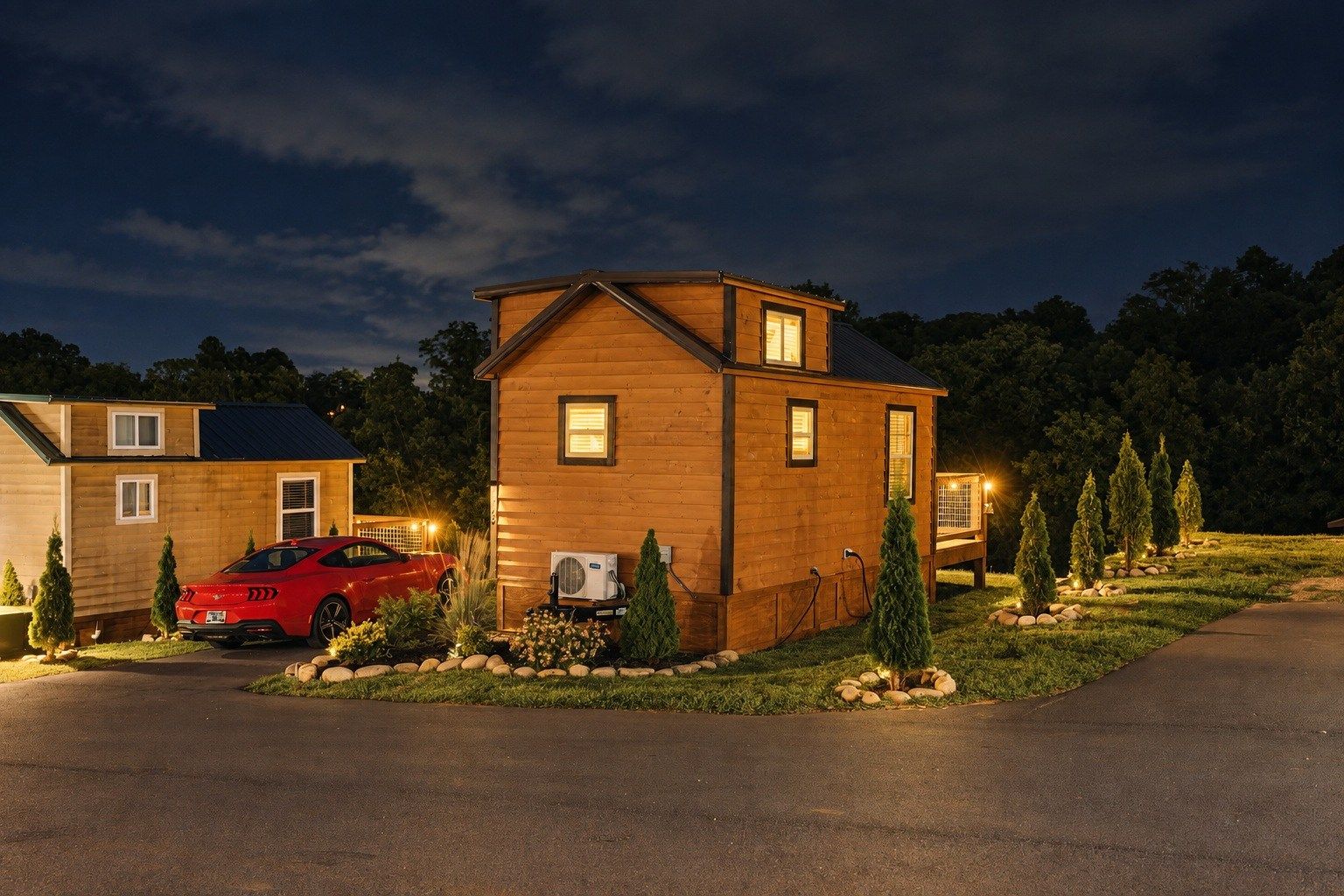 A two-story wooden cabin with a red sports car parked in front, surrounded by a landscaped garden with small trees and a paved driveway.