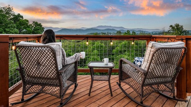 A deck with wicker rocking chairs and a small table, overlooking a scenic landscape with mountains and trees. - The Lookout — Romantic Tiny Home w/ Mountain Views (Sevierville)