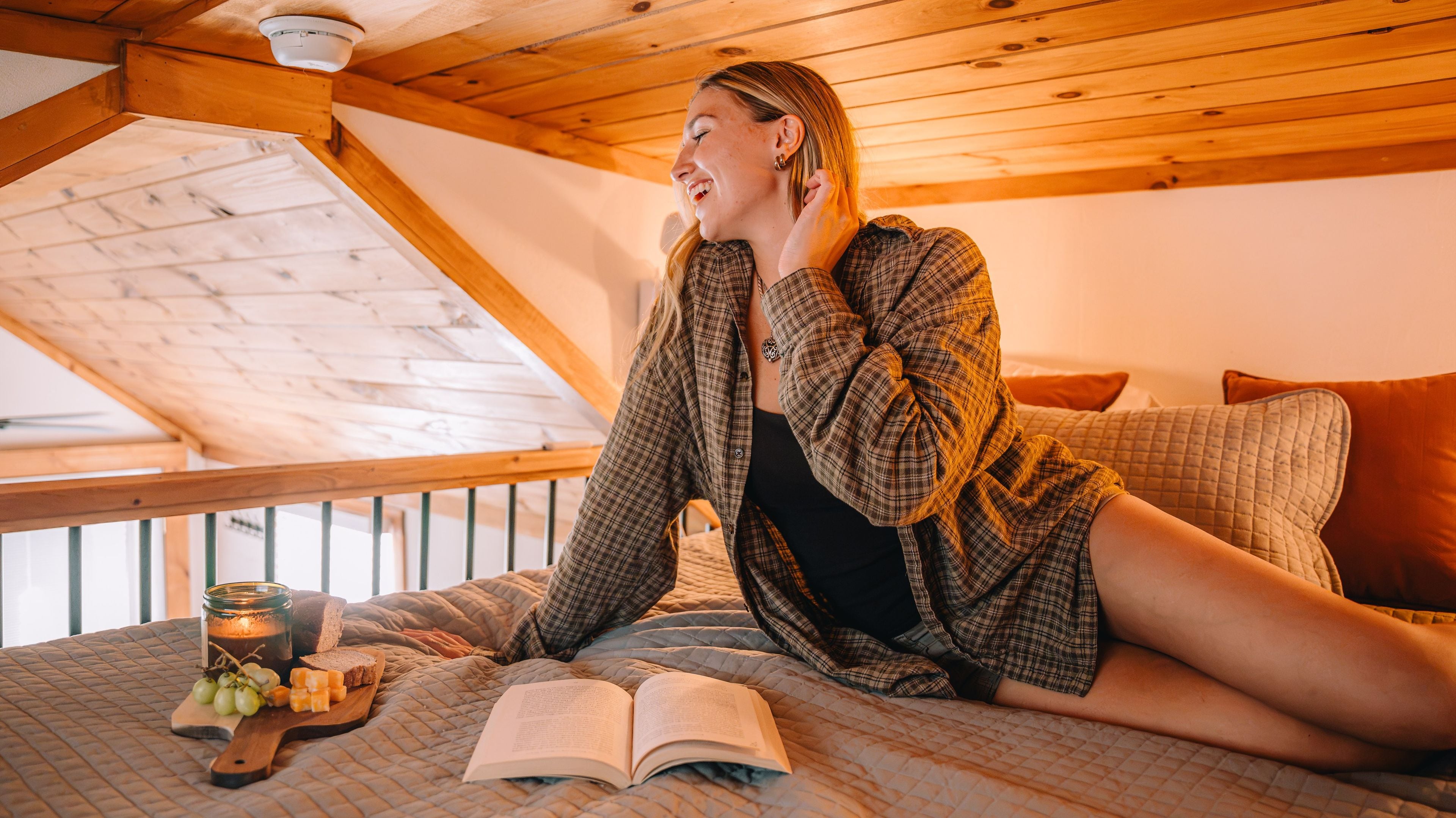 A person sitting on a bed in a room with a sloped ceiling and wooden beams, reading a book.
