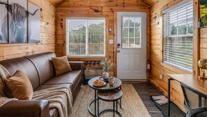 A cozy cabin interior with a wooden ceiling, a leather sofa, a small round table with a plate of food, and a door with a window. - The Lookout — Romantic Tiny Home w/ Mountain Views (Sevierville)