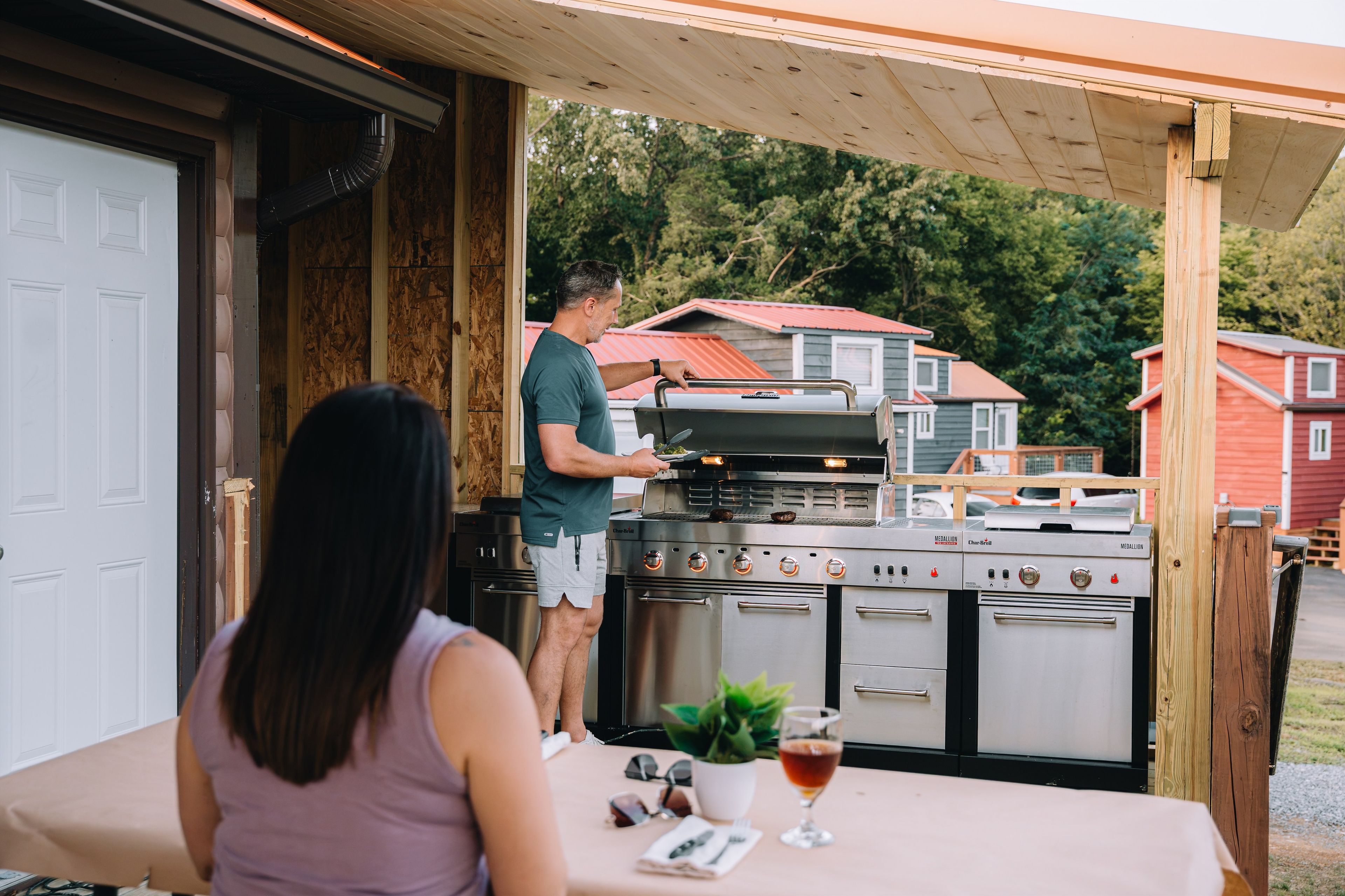 A person grilling outdoors with a large grill and a table with a glass of drink and a plant.