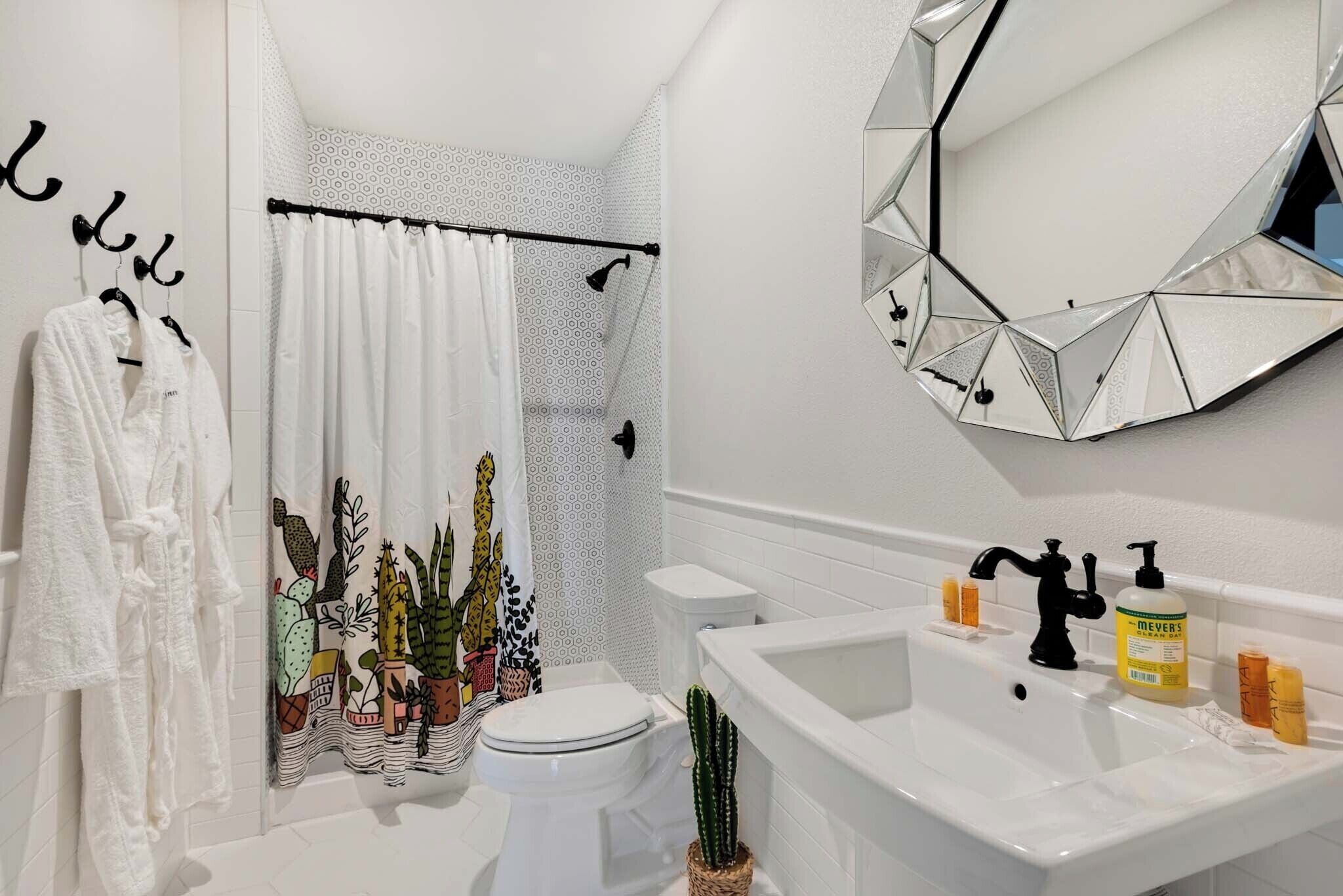 A bathroom with a white sink, a toilet, a shower curtain with a cactus pattern, and a mirror with a geometric frame.