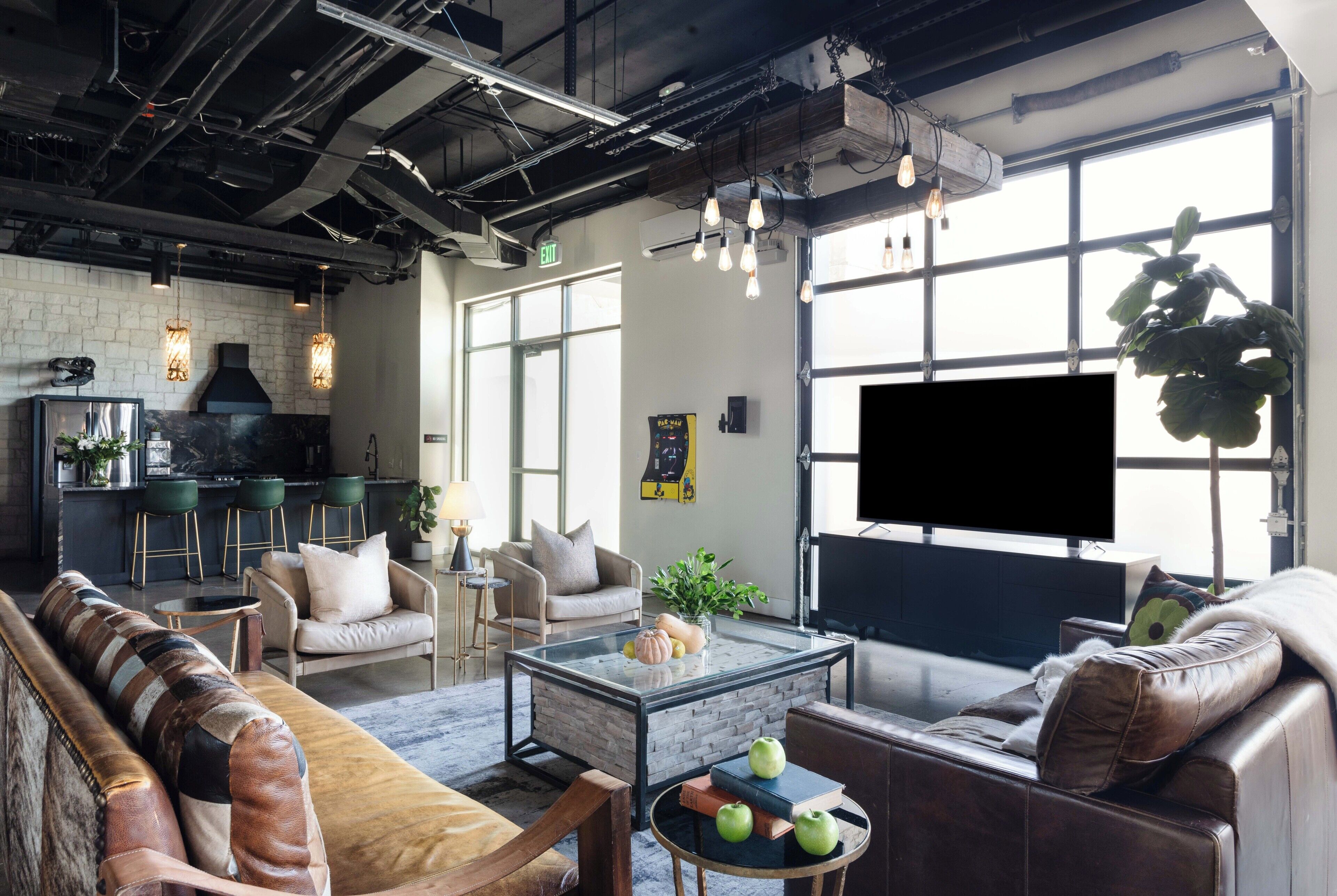 A modern living room with a sofa, armchair, and coffee table, a kitchen area with bar stools, and a large window with a TV.