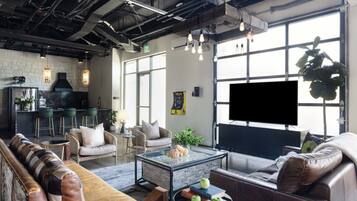 A modern living room with a sofa, armchair, and coffee table, a kitchen area with bar stools, and a large window with a TV.