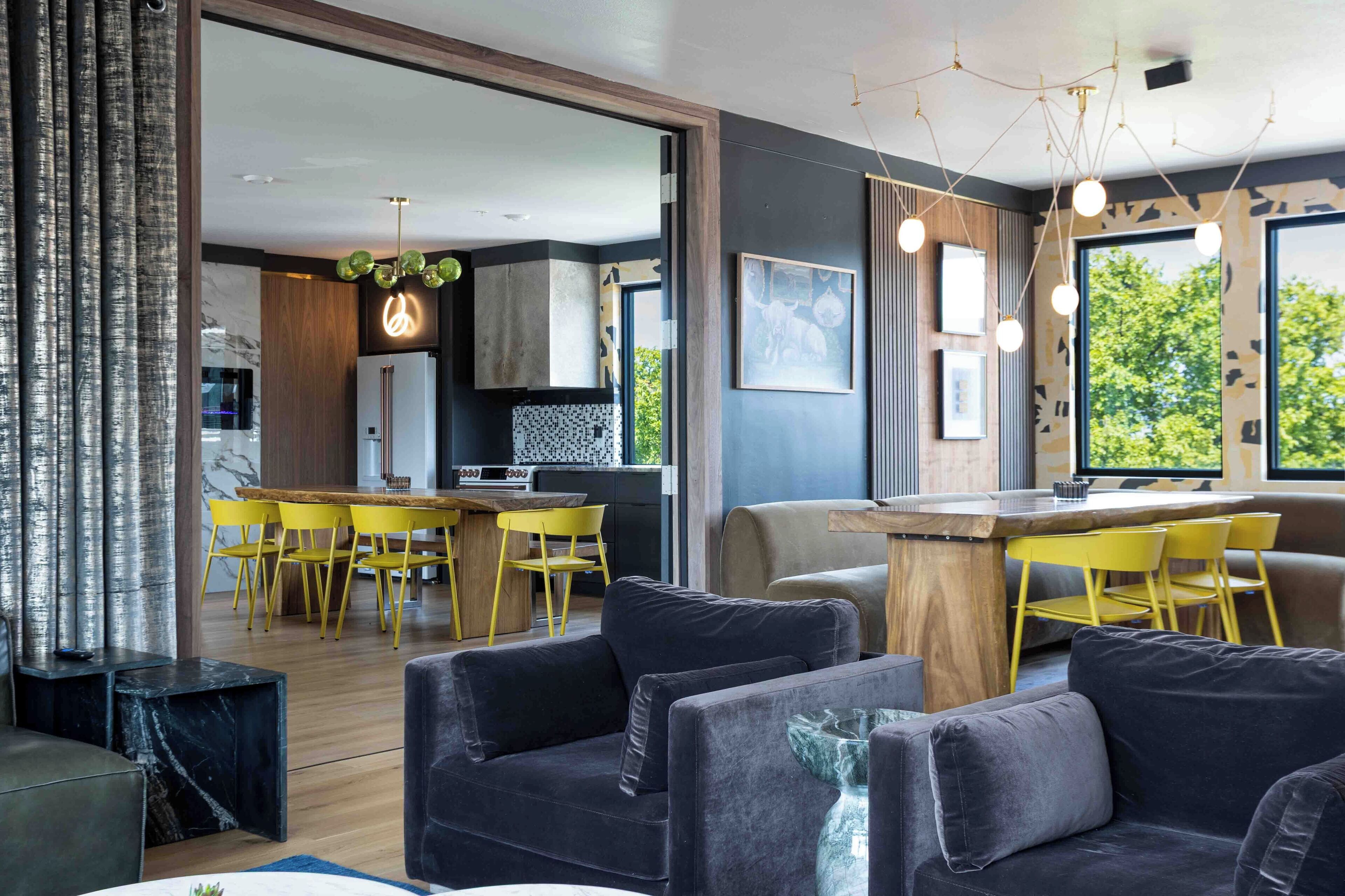 Un salón moderno con un sofá oscuro, una mesa de centro de madera y sillas amarillas. Se ve en el fondo una cocina con encimera de madera y una zona de comedor con sillas amarillas.