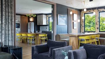 Un salón moderno con un sofá oscuro, una mesa de centro de madera y sillas amarillas. Se ve en el fondo una cocina con encimera de madera y una zona de comedor con sillas amarillas.