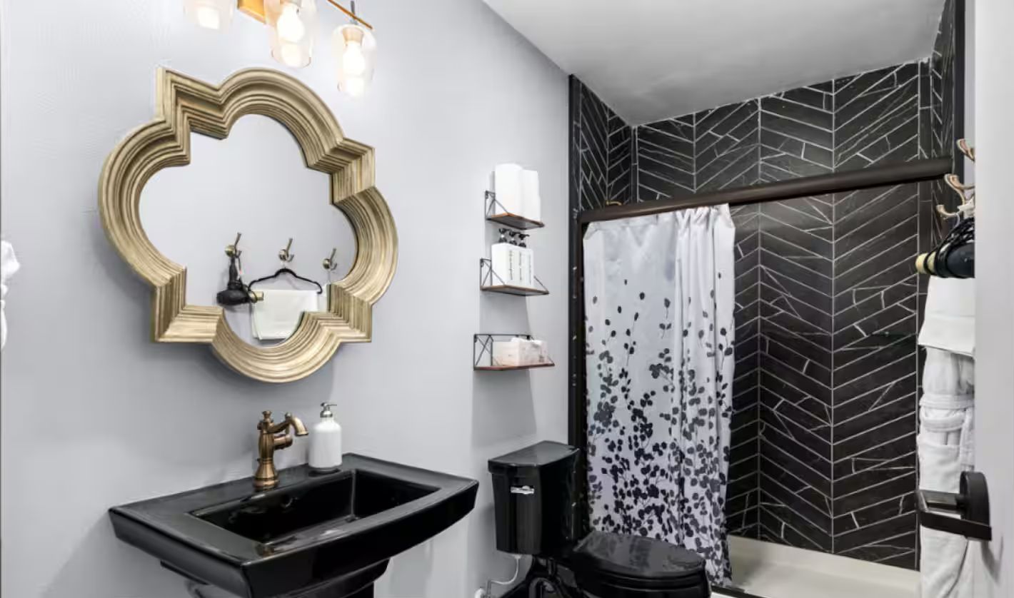 Un baño moderno con un lavamanos negro, espejo con marco dorado y una ducha con cortina de hojas en blanco y negro.