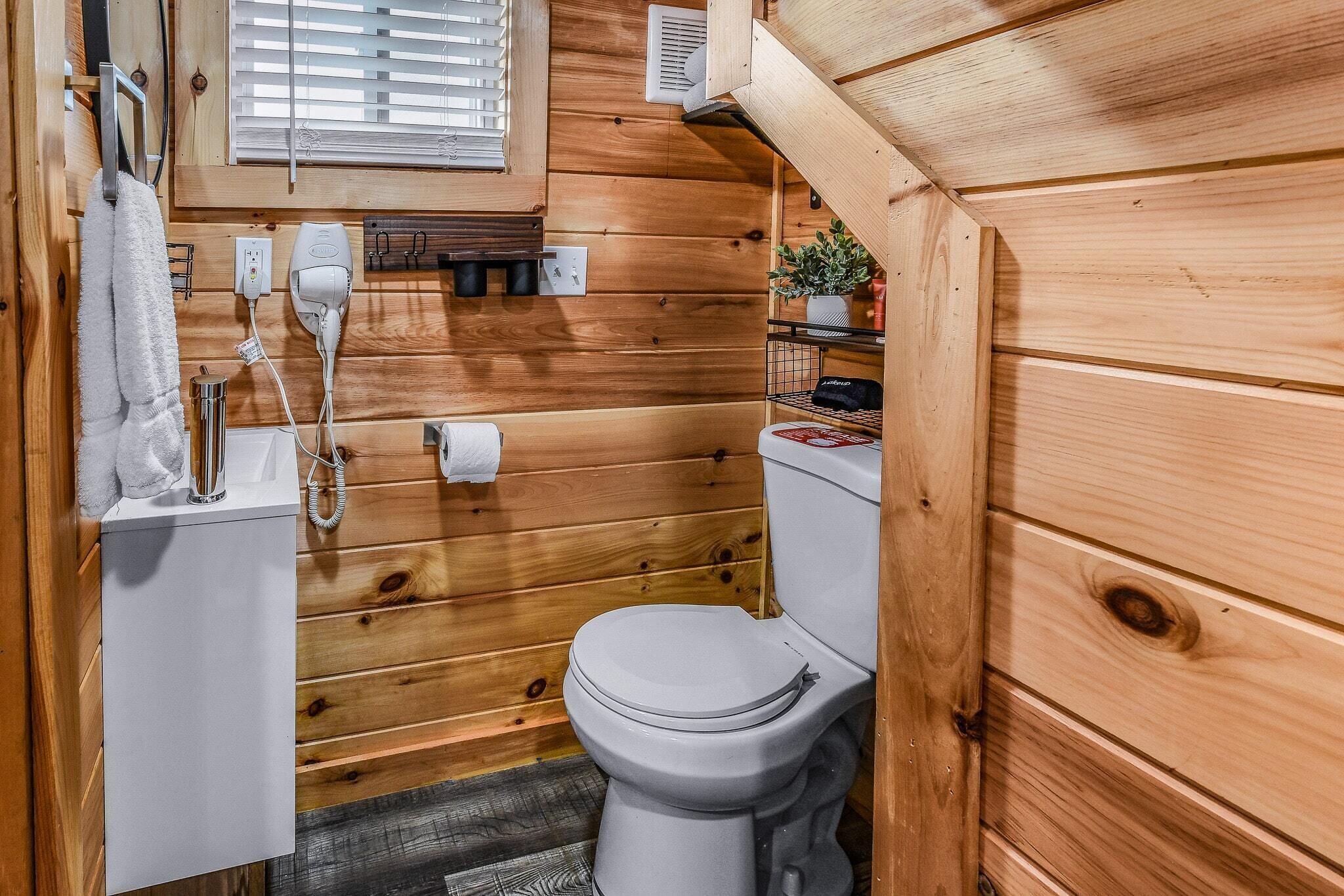 A compact wooden bathroom with a toilet, a towel rack, a hairdryer, and a small shelf with toiletries.