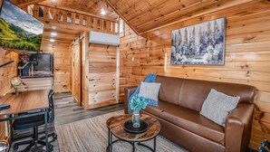 Interior d'una cabana de fusta amb un sofà de cuir marró, una taula de centre rodona amb un ram, un escriptori amb un globus i un quadre d'una escena forestal.