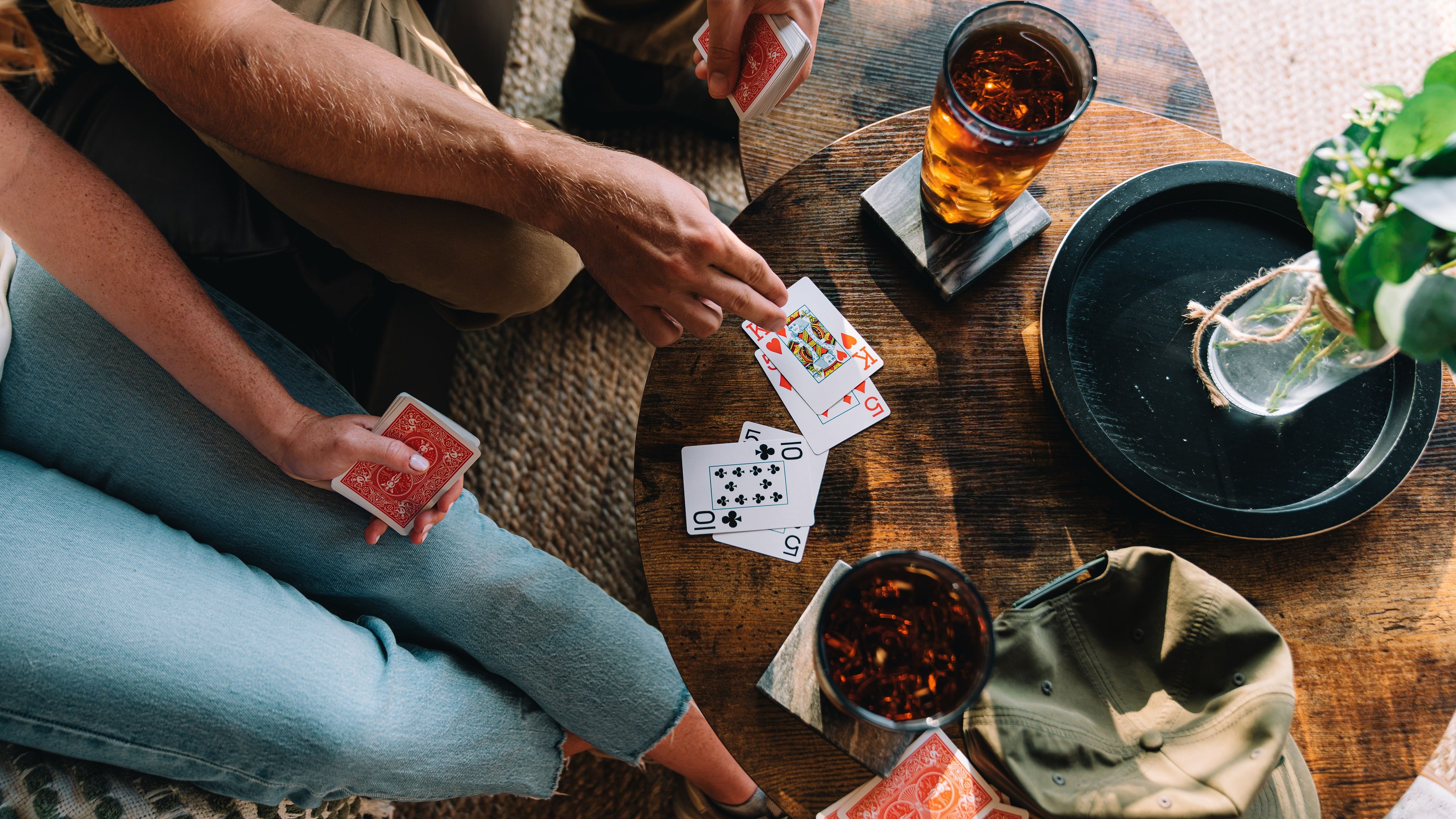 Une personne tenant un jeu de cartes et un verre d’une boisson sombre sur une table en bois.