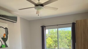 Un dormitorio con cama, ventilador de techo, ventana con cortinas y vistas al exterior.