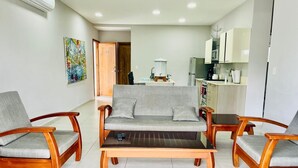 Una sala de estar con un sofá y dos sillones, una mesita de centro, un cuadro en la pared y una cocina al fondo.