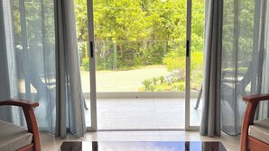 Habitación con una mesa de vidrio, dos sillas de madera y vistas a un balcón con vegetación a través de una puerta corrediza de vidrio.