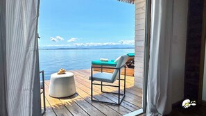 Une terrasse en bois avec une chaise et une petite table ronde, donnant sur une étendue d’eau calme et une terre lointaine.