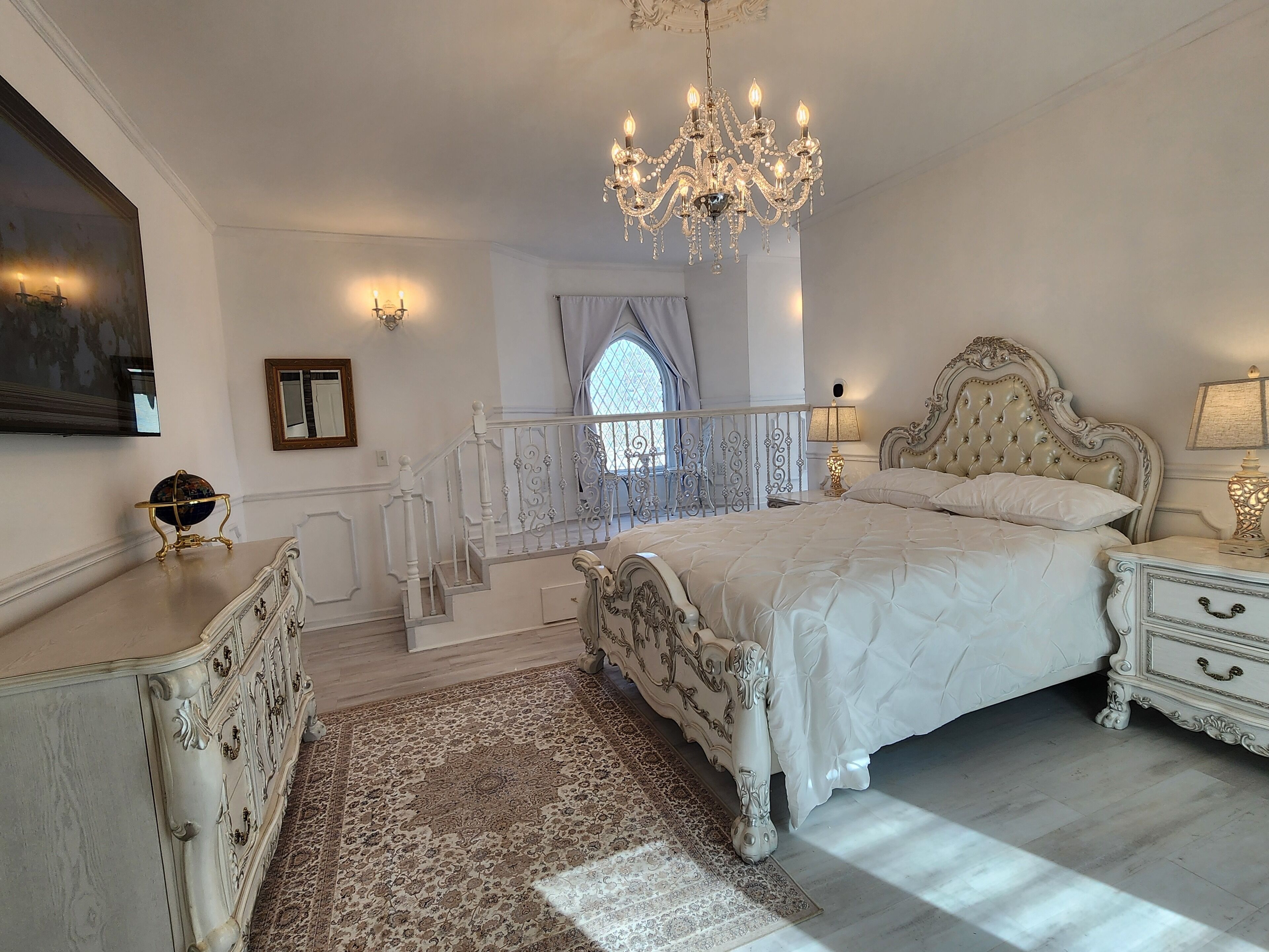 A bedroom with a large bed, ornate white furniture, a chandelier, and a view through a window.