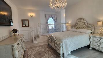 A bedroom with a large bed, ornate white furniture, a chandelier, and a view through a window.