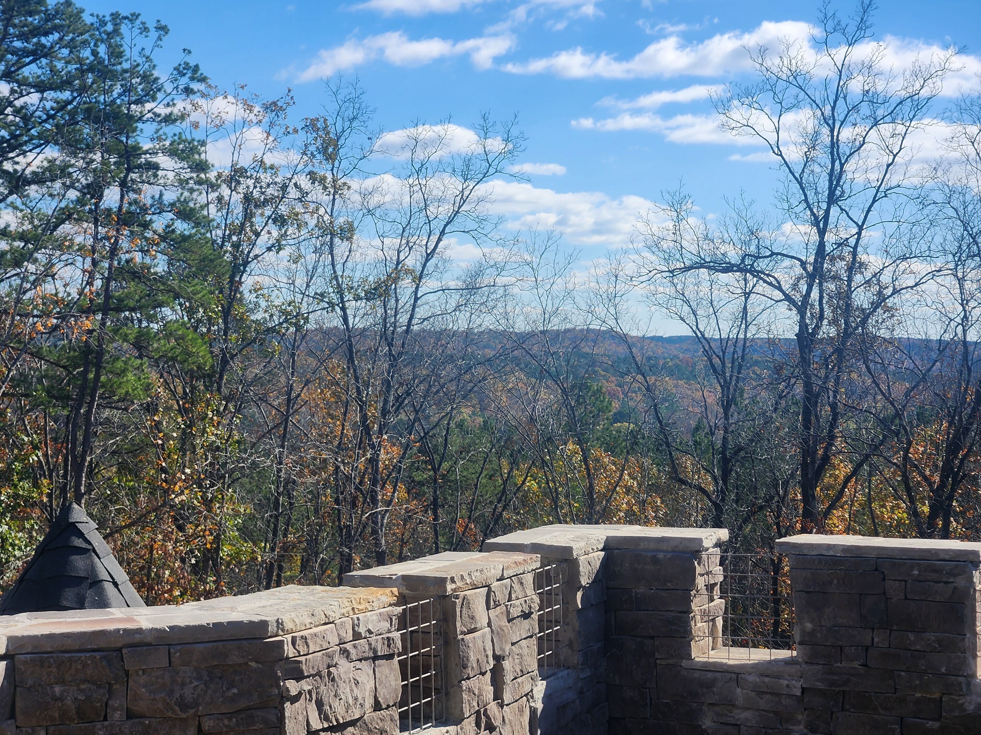 A stone wall with a metal railing, overlooking a forested area with autumn-colored trees.