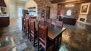 Un vaste espace dédié à la salle à manger, avec une grande table et des chaises en bois, une cheminée en pierre et une cuisine à l’arrière-plan.