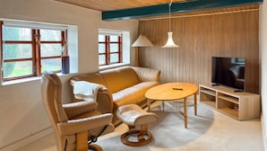 A cozy living room with a sofa, armchair, ottoman, and a small round table. There is a television on a wooden stand and a window with a view of greenery. - Lovely home in Haderslev with WiFi (Haderslev)