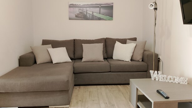 A modern living room with a sectional sofa, a small table, and a painting of a dock. - Patisia Metro Station 2 bdrm apartment by MPS (Athina)