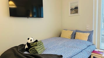 Un dormitorio con una cama, un televisor montado en la pared, una ventana con persianas y una pequeña mesa con un libro.