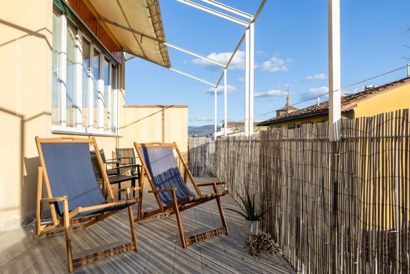A patio with two wooden chairs and a table, a wooden fence, and a building with a yellow facade.