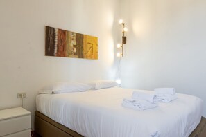 A hotel room with a bed, white bedding, a wall-mounted light fixture, and a painting.