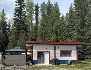 A small cabin with a red roof, a covered outdoor seating area, and a view of dense forest.