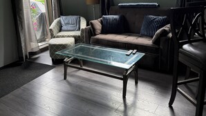 Una sala de estar con una mesa de centro de vidrio, un sofá y un sillón. Hay una ventana con cortinas, una lámpara y un cuadro en la pared.