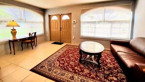 A living room with a brown leather sofa, a round coffee table, a wooden side table with a lamp, two doors, and large windows with blinds. - Spacious & Expansive Miami Home With Free Parking (Homestead)