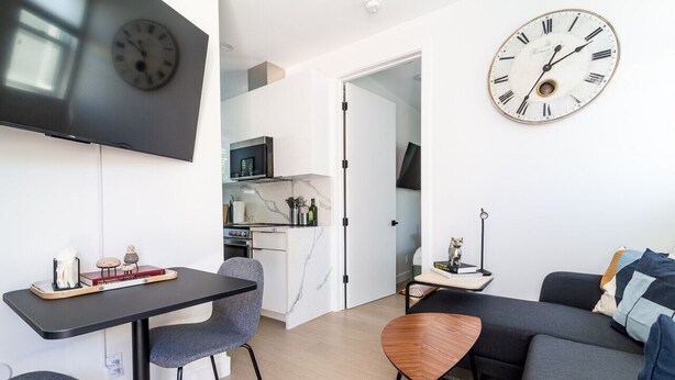 A modern living room with a sofa, a dining table with chairs, a TV mounted on the wall, and a kitchen area visible through an open door.