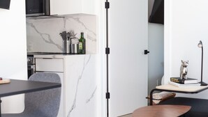 A compact kitchen with a marble backsplash, a dining area with a table and chairs, and a small side table with a lamp and a book.