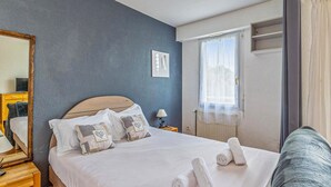 A neatly made bed with white linens and decorative pillows, a bedside table with a lamp, and a window with sheer curtains. - Magnifique app à la Rochelle (La Rochelle)