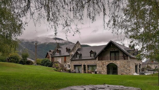 A stone house with a sloped roof, surrounded by a grassy area and trees.