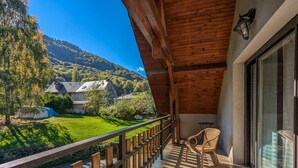 A balcony with a view of a mountain and a stone building. There are two wicker chairs and a table on the balcony.