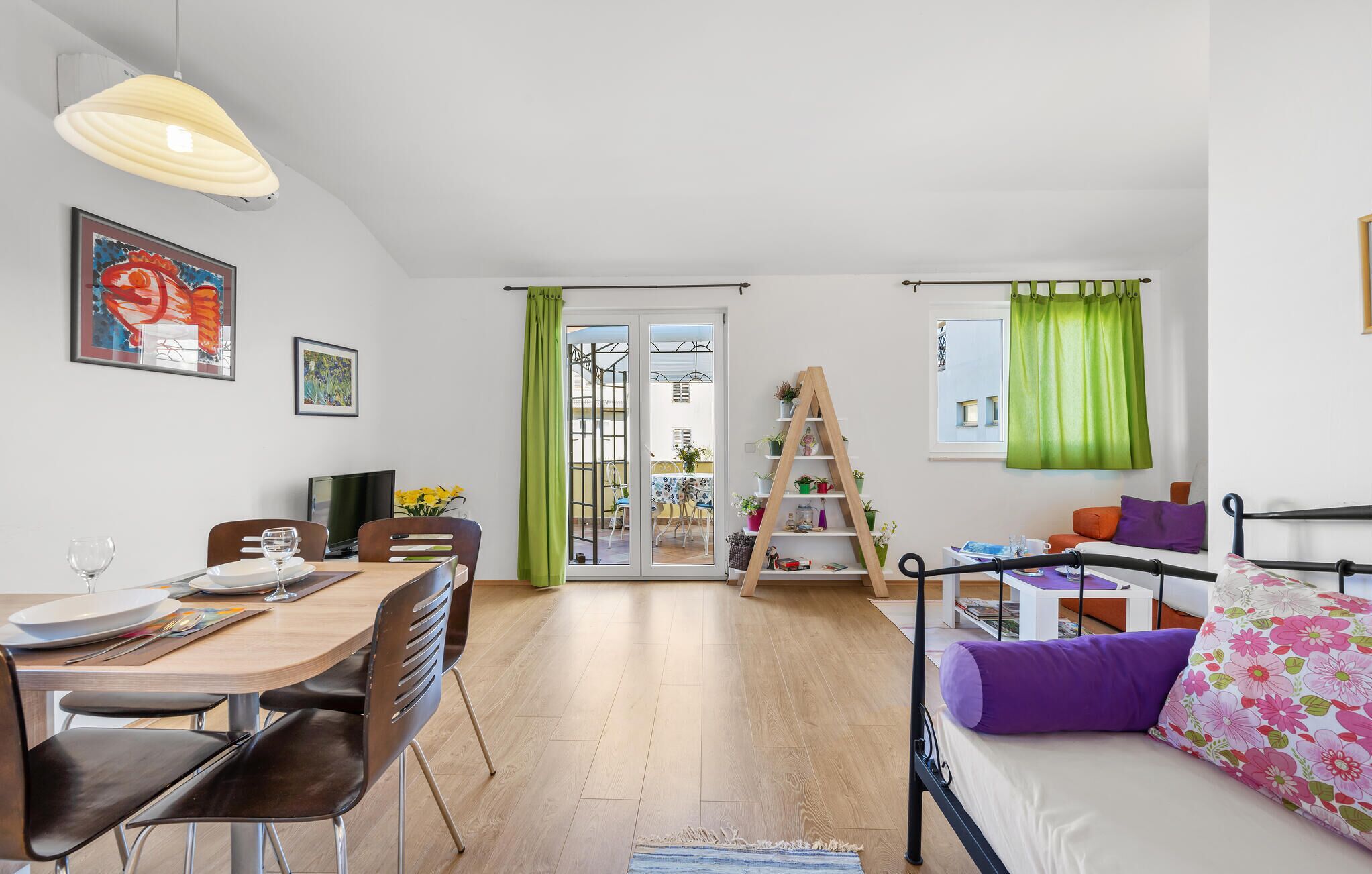 Ein modernes Wohnzimmer mit Essbereich, einem Bett mit floralem Bettzeug und einem Stegregal mit Pflanzen.